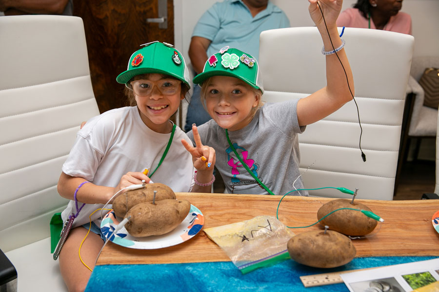 Two girls with green baseball hats and seated at a table with potatoes.