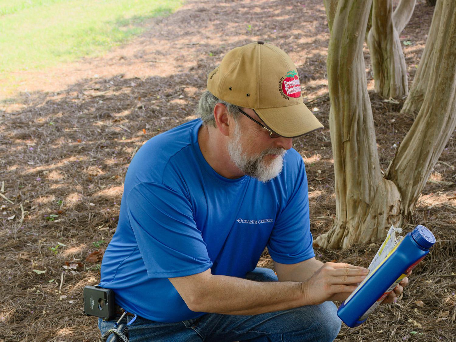 A man kneels on the ground beside a pump sprayer to read the label on the back of a pesticide jar.