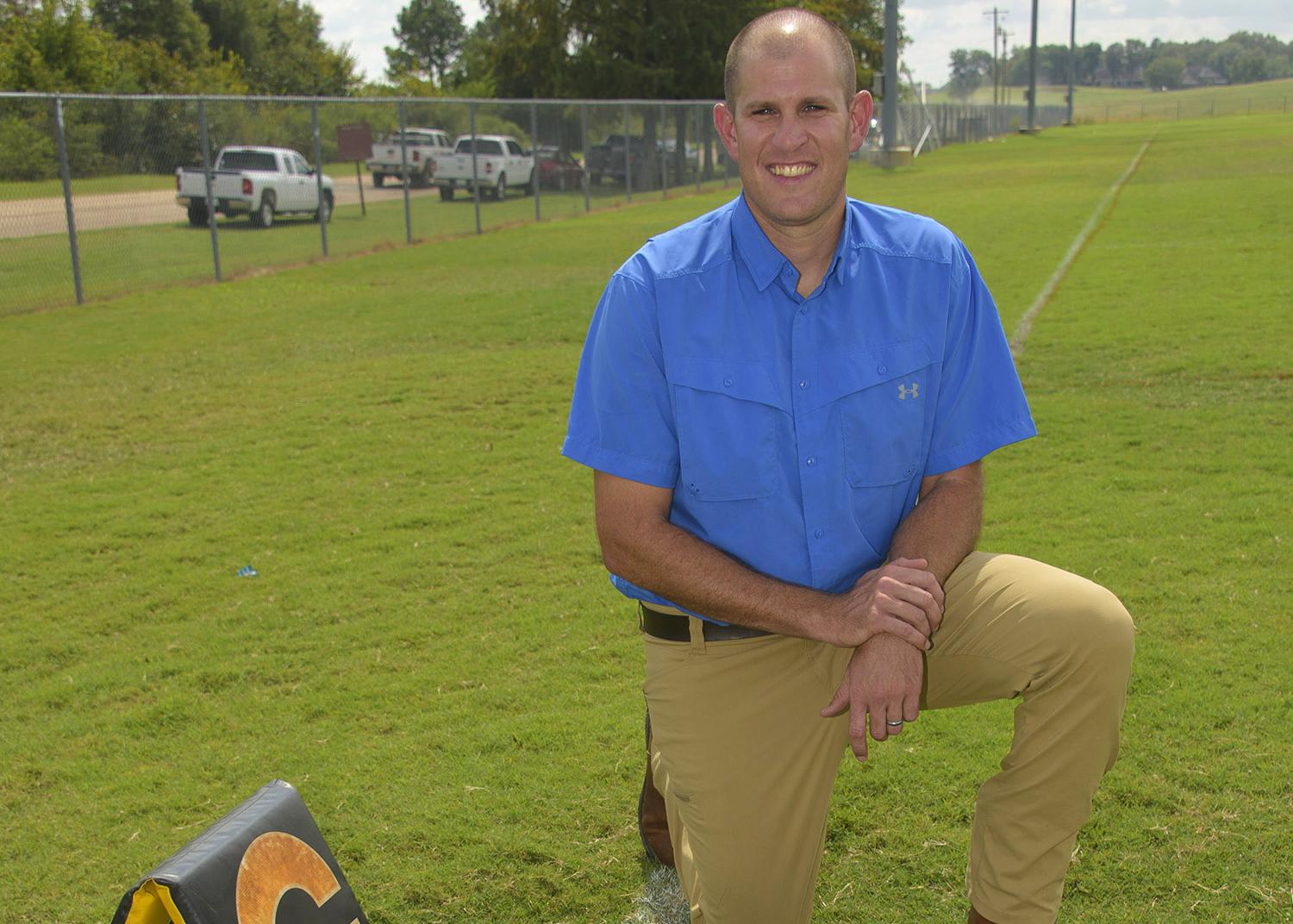 A man kneels on a football practice field beside a football.