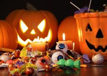 Orange pumpkins and a carved jack-o-lantern sit on a table behind a smiling ceramic jack-o-lantern candy dish. Toys, candy and candles are displayed in front of the pumpkins.