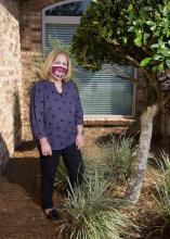 A woman wearing a mask stands in the landscape outside a house.