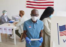 Voter wearing mask, standing by booth and looking at camera.