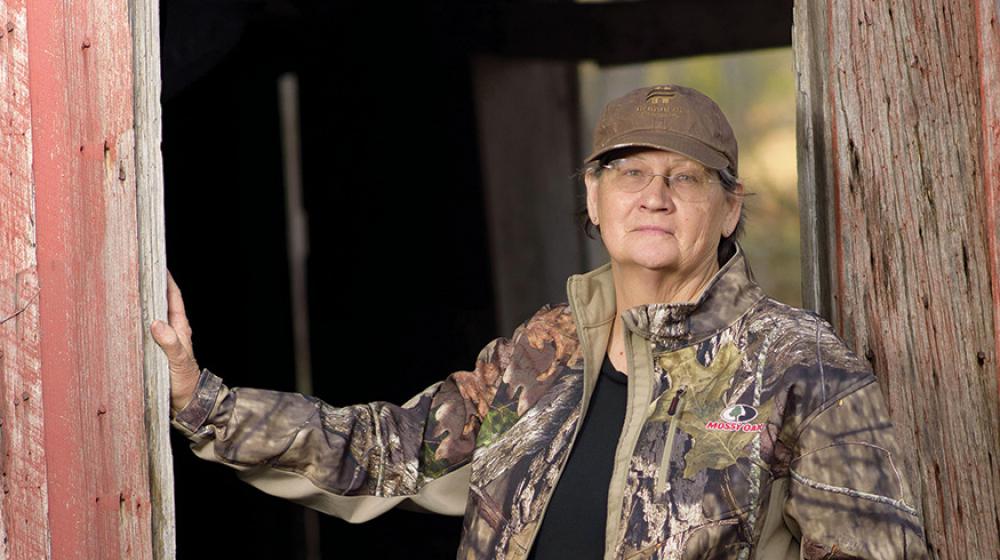 A woman wearing a camouflage jacket and pants rests her hand on the entrance to an old wooden building. 