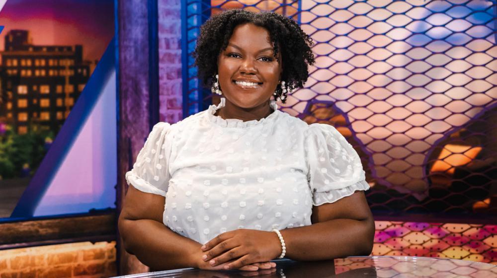 A smiling woman wearing a white shirt sits behind a desk with her hands resting on the desk in front of her.