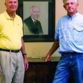 Two men, one in a yellow polo and the other in blue, stand in front of a portrait of a man