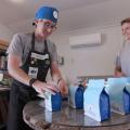 : A man with a blue cap and glasses grabbing a blue bag of coffee for a blonde, smiling woman.