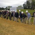 A group of more than a dozen people in hard hats break ground with shovels.