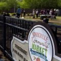 Shoppers can be seen browsing in the background of a farmers market sign.