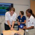 One teenager clasps both of her hands to press on a training manikin’s chest, while another female student holds a ventilator mask over its face. A nurse in the background watches a large wall-mounted monitor.