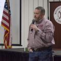 Man speaks into a handheld microphone. Background includes the Mississippi State University seal and an American flag.