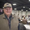 A man in a baseball cap stands in front of long tables with a crowd of people in the background.