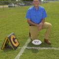 A man kneels on a football practice field beside a football.