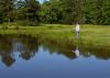 Older man holds a fishing pole on the shoreline while watching a red cork on a quiet pond.