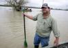 A man in work clothes, baseball cap and wading boots stands in water outside his boat holding a paddle.