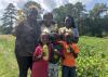 Two women and four children stand in a vegetable garden while holding yellow squash.