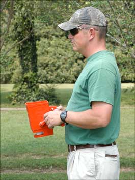 A man stands on a green lawn while holding a small hand-powered red spreader used for spreading fire ant bait.